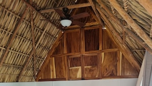 A cozy bedroom with a sloped ceiling, wooden beams, a bed with a headboard, a nightstand, a fan, and a wooden ladder.