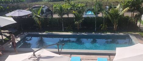 A swimming pool with lounge chairs and umbrellas, surrounded by tropical vegetation and thatched-roof structures.