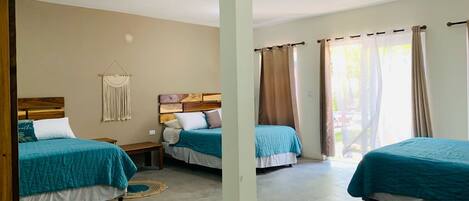 A hotel room with three beds, a macrame wall hanging, and a large window with curtains.