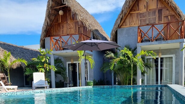 A clear swimming pool with a tiled edge, surrounded by two thatched-roof buildings and palm trees.