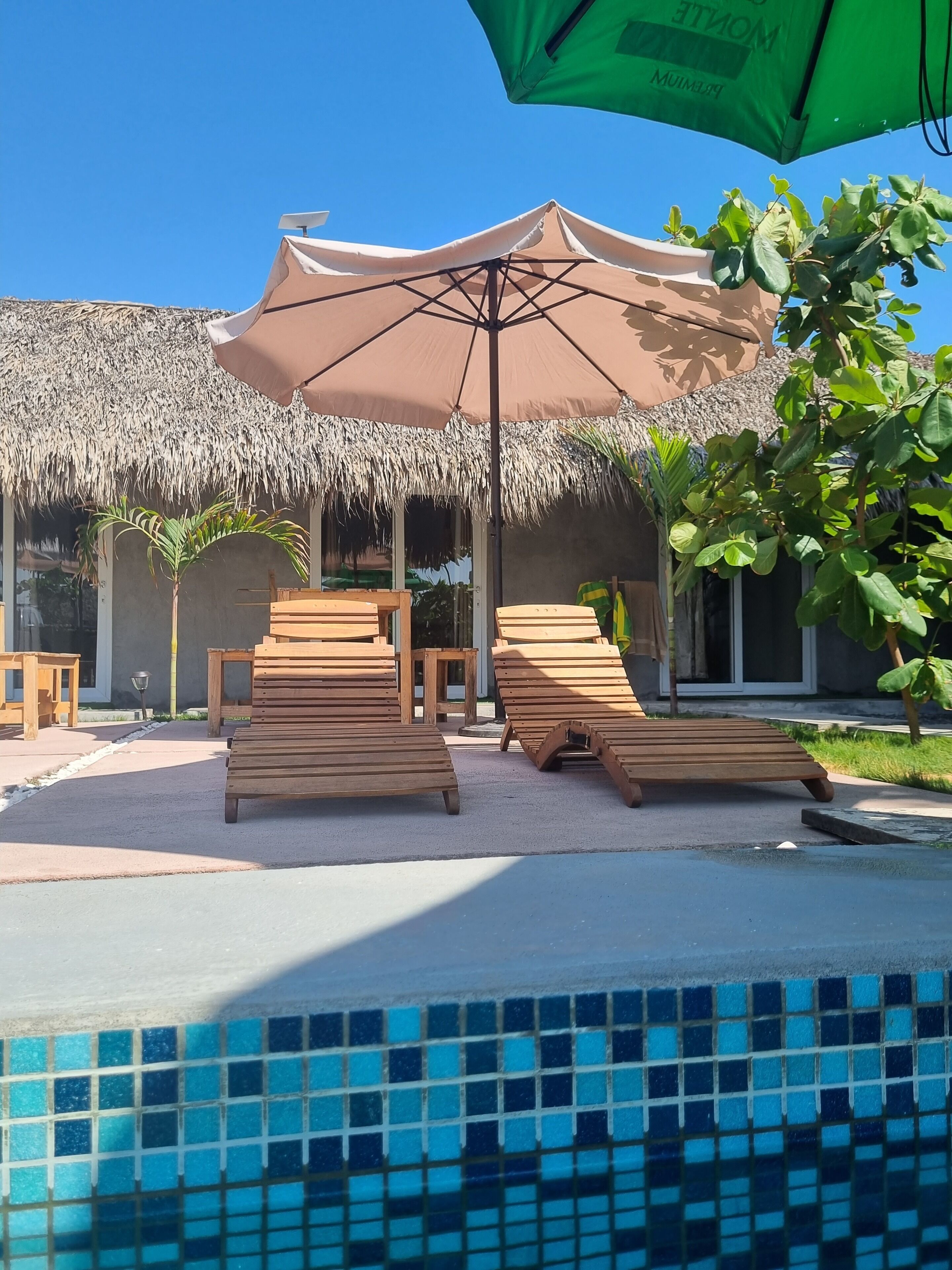 A poolside area with wooden loungers, a thatched-roof structure, and a green umbrella.