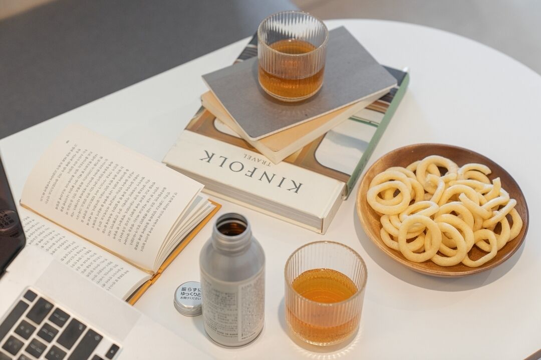 Una mesa con una computadora portátil, un libro abierto, una botella, un vaso y un bol de aperitivos en forma de aros.
