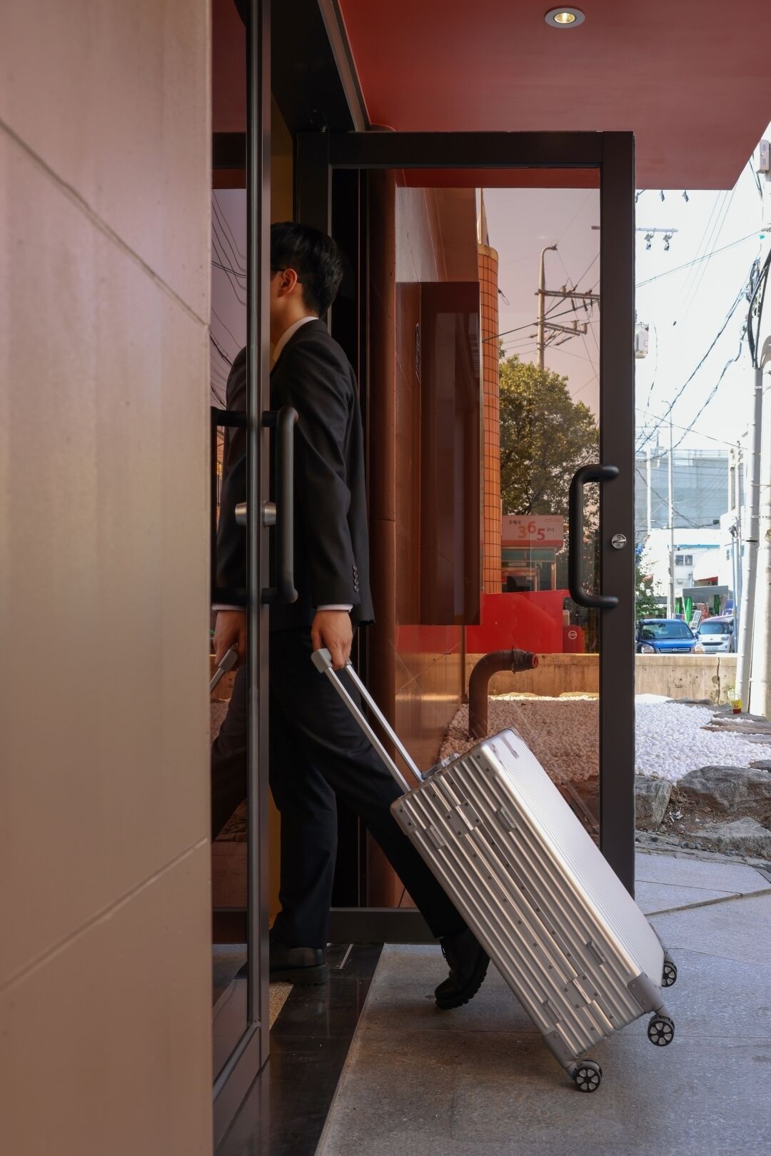 A man exiting a building with a wheeled suitcase.