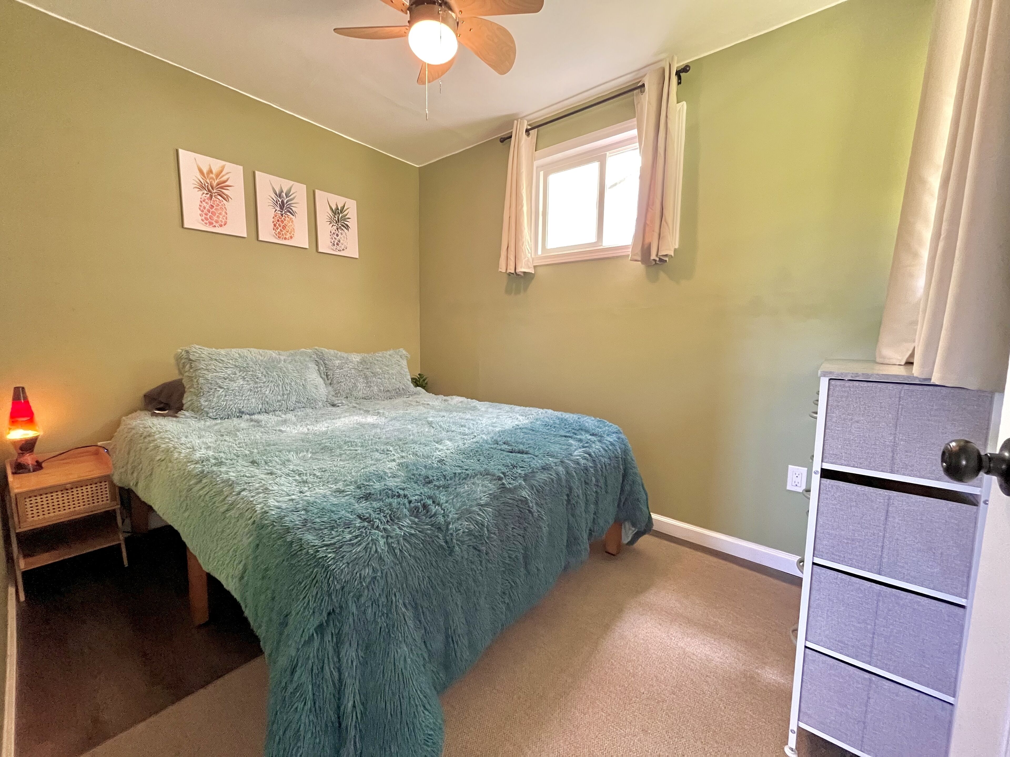 A bedroom with a bed, a nightstand with a lamp, a ceiling fan, and a window with curtains.