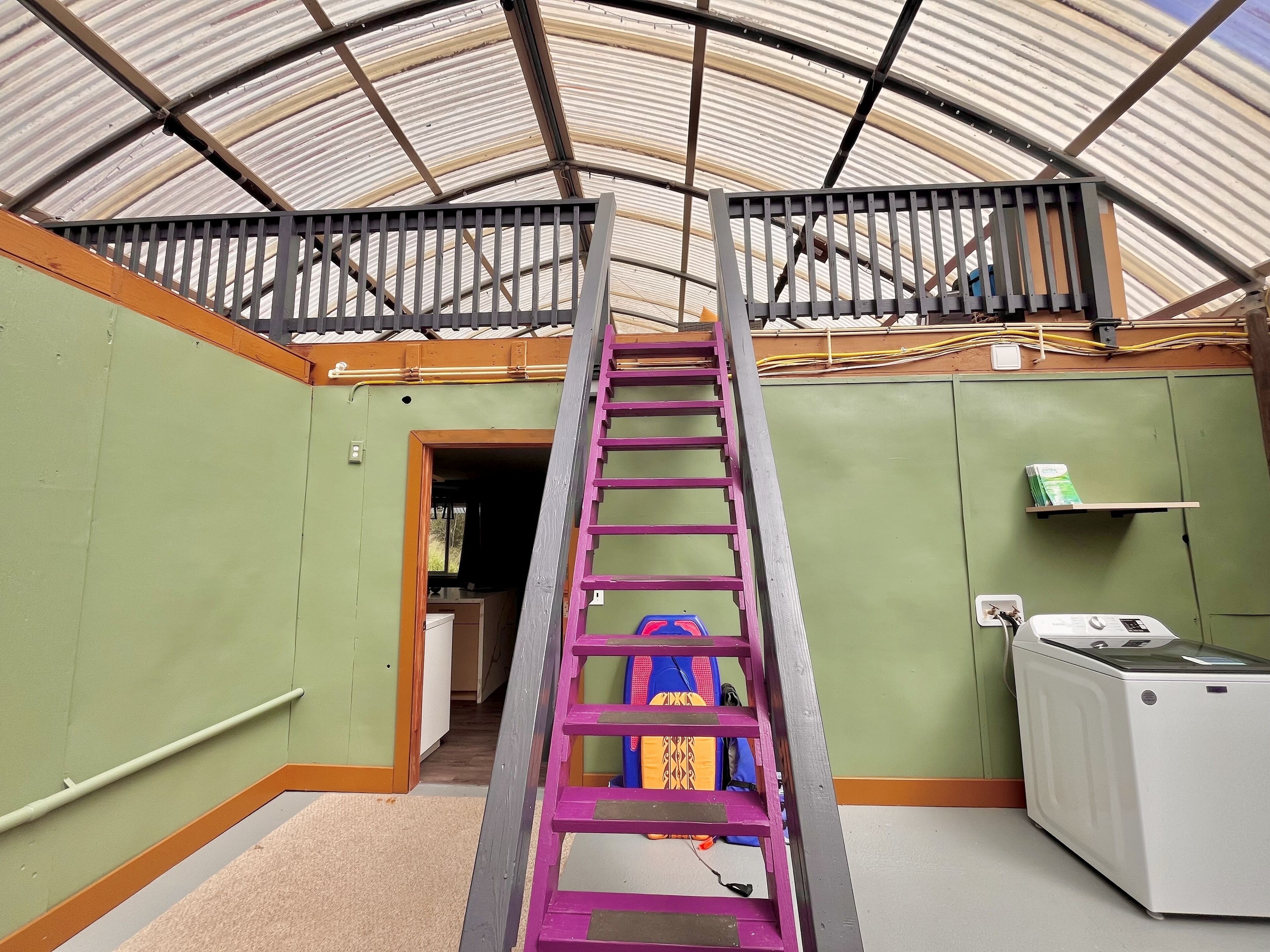Una habitación con una escalera metálica que lleva a un balcón, una lavadora y un estante con objetos en la pared.