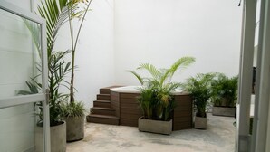 Un petit espace extérieur clos, doté d’un sol en béton, entouré de murs blancs et agrémenté d’un banc en bois, de plantes en pot et d’un petit coin salon.
