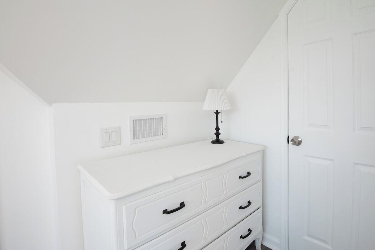 A white dresser with drawers, a lamp, a vent, and a door in a room with white walls.