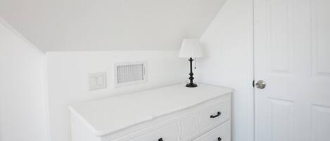 A white dresser with drawers, a lamp, a vent, and a door in a room with white walls.