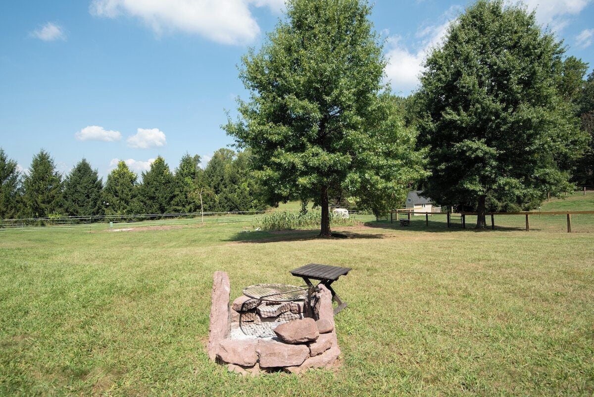 A stone fire pit with a table and bench in a grassy field with trees in the background.