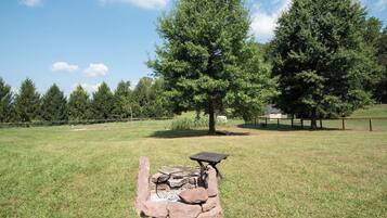 A stone fire pit with a table and bench in a grassy field with trees in the background.