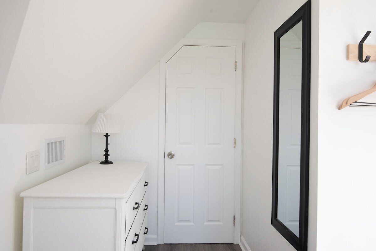 Une commode blanche avec des tiroirs, une lampe, une porte blanche et un miroir au mur.