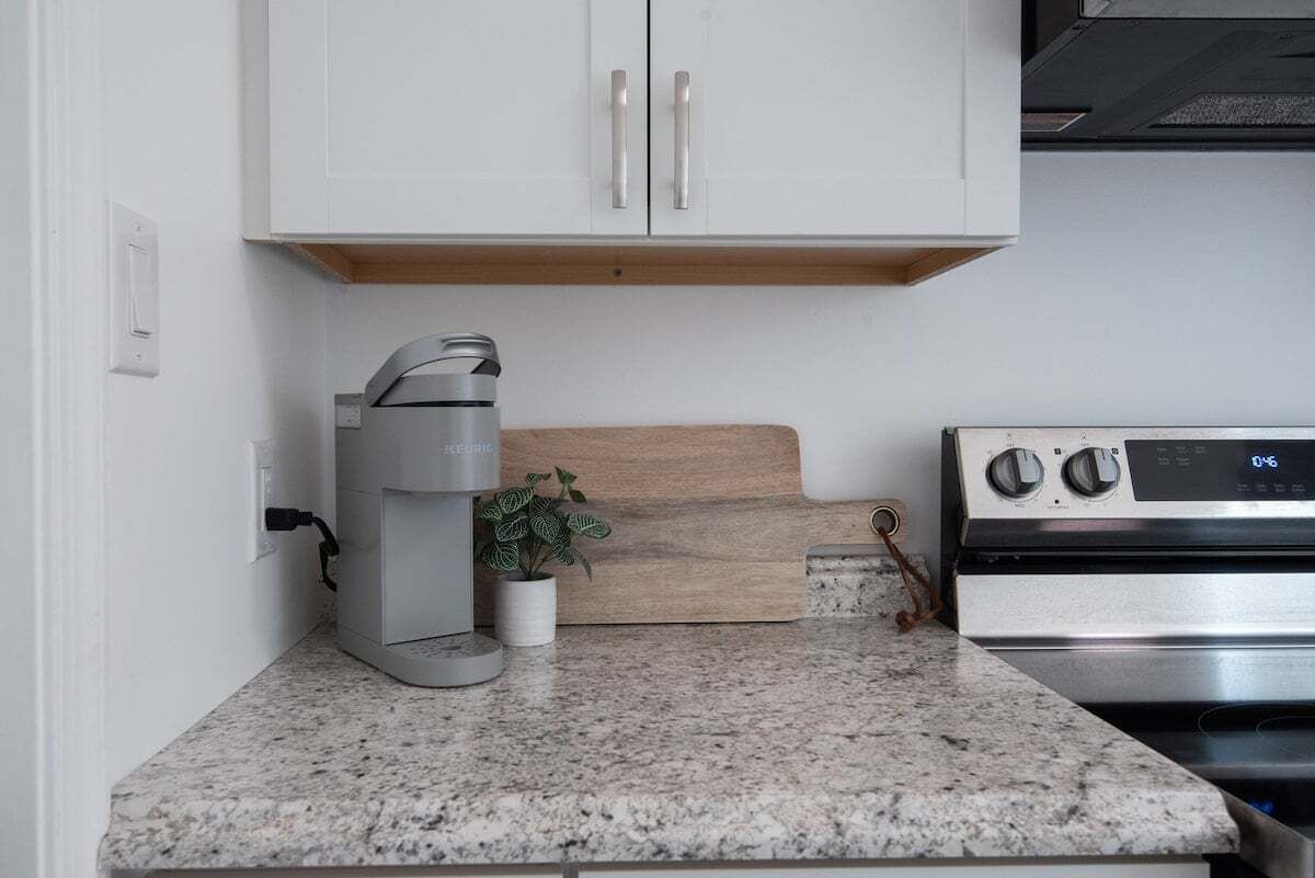 A kitchen with white cabinets, a stainless steel stove, a coffee maker, a wooden cutting board, and a potted plant.