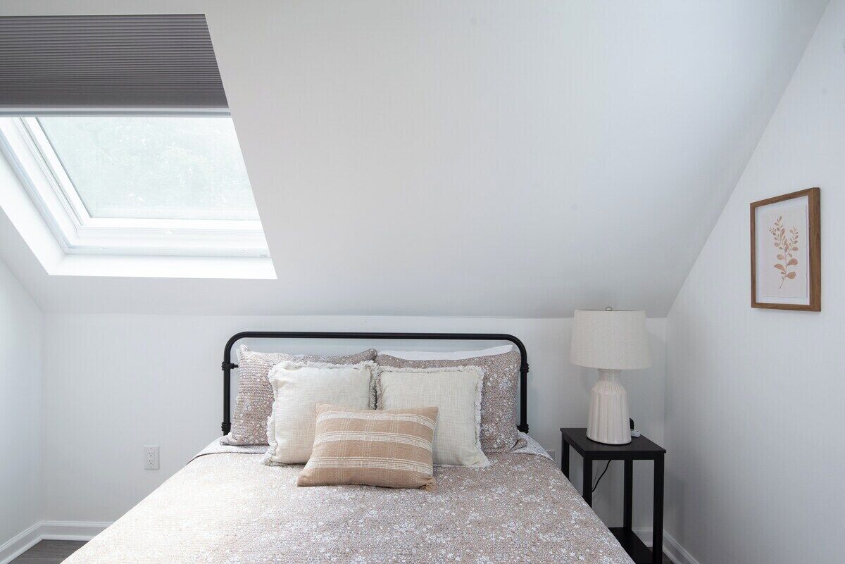 A bedroom with a skylight, a bed with pillows, a bedside table with a lamp, and a framed picture on the wall.
