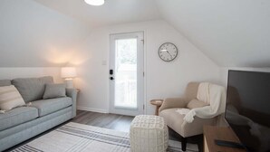 Ein Wohnzimmer mit grauem Sofa, einem beigen Sessel, einem Fernseher, einer Uhr und einer Tür mit Jalousien.