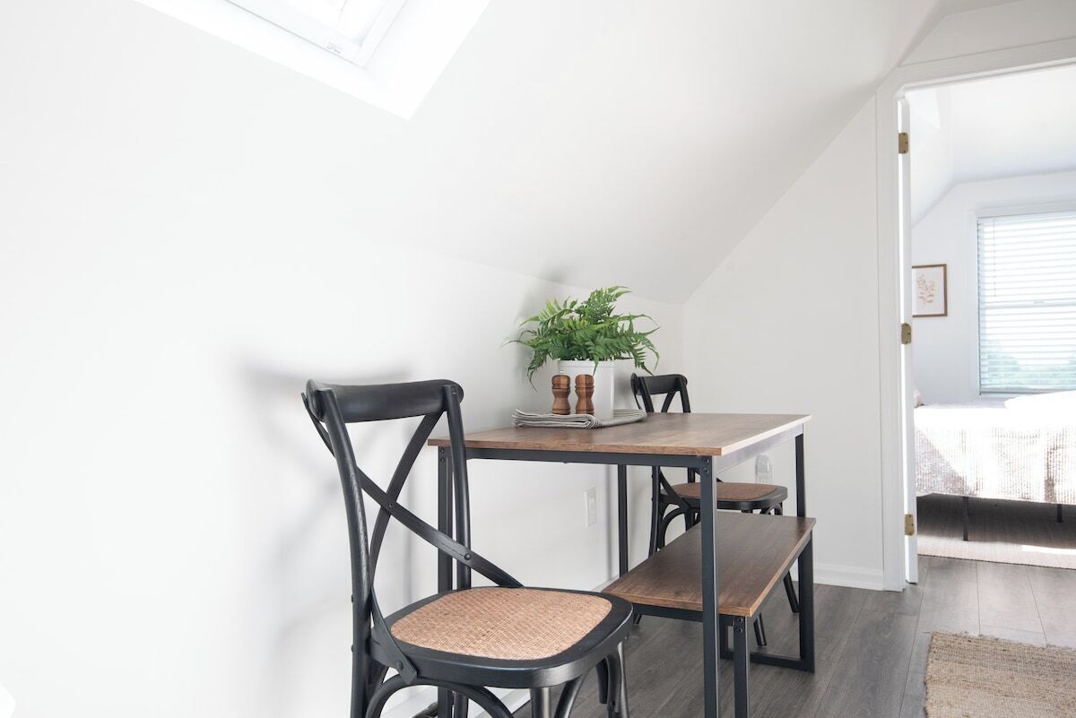 Uma área de refeições com mesa e cadeiras, uma planta sobre a mesa, e vista para o exterior através de uma janela.