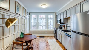 Une cuisine avec un parquet en bois disposé en chevrons, une table à manger avec des chaises, un réfrigérateur et une fenêtre ornée de barreaux décoratifs.