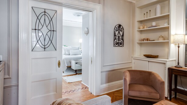 Une pièce bien éclairée avec une porte blanche, un fauteuil beige et une table en bois. On y trouve une étagère blanche ornée d’objets décoratifs et une lampe.