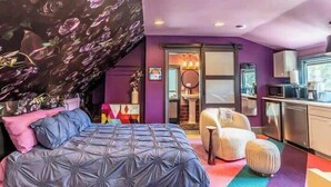 Quarto com cama, uma cadeira, mesa pequena e área de cozinha compacta.
