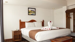 Una cama bien hecha con cabecera de madera, una mesita de noche con un teléfono y un banco de madera.