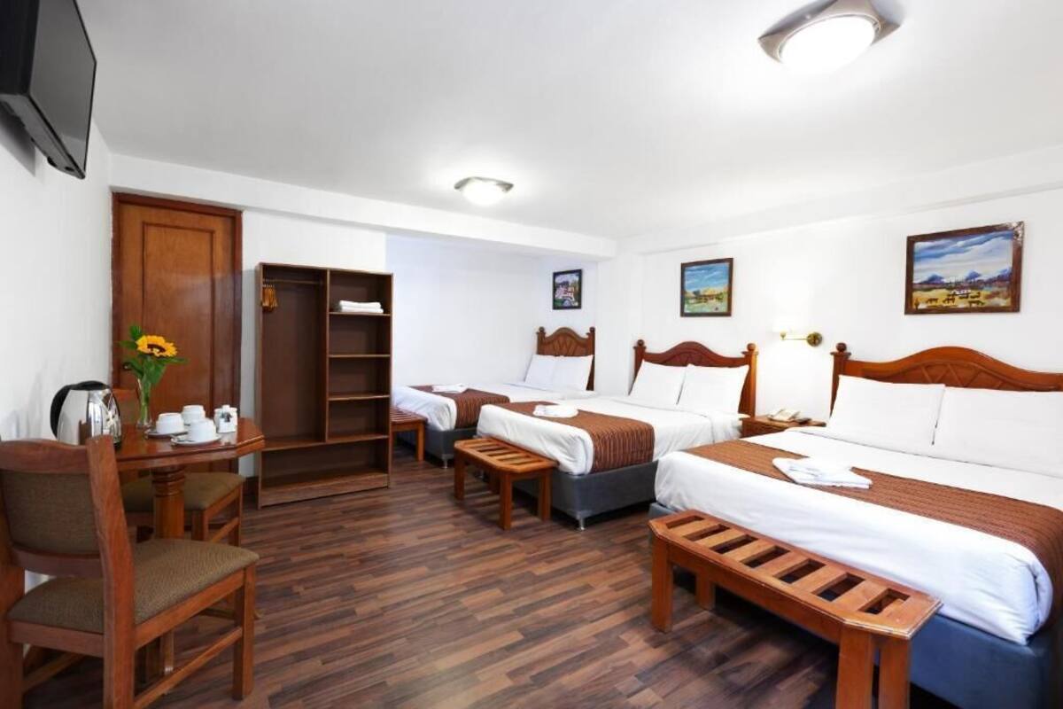 A hotel room with two beds, a wooden desk with a chair, a small table, and a TV mounted on the wall.