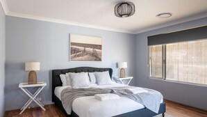 A modern bedroom with a dark headboard, white bedding, and a large window with blinds. - Spacious 4x3 home, just minutes away from the Margaret River main street. (Margaret River)