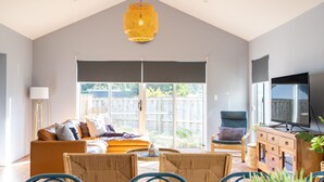 A spacious living room with a high ceiling, a large window, a sofa, a dining table, and a television. - Spacious 4x3 home, just minutes away from the Margaret River main street. (Margaret River)