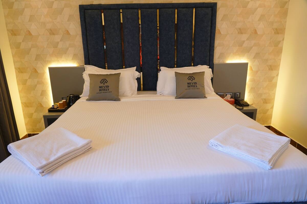 A hotel room with a large bed, two pillows with the hotel's logo, a nightstand with a phone and a lamp, and a geometric-patterned wall.