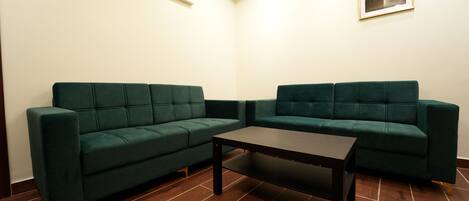 Una habitación con dos sofás verdes, una mesa de centro de madera oscura, suelo de madera, una luz empotrada en el techo y un cuadro en la pared.