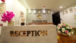 A reception desk with the sign "RECEPTION" and "MEVID HOTELS LUXURY REDefined". There are clocks on the wall, a vase with flowers, and framed pictures. - Mevid hotels (Hyderabad)