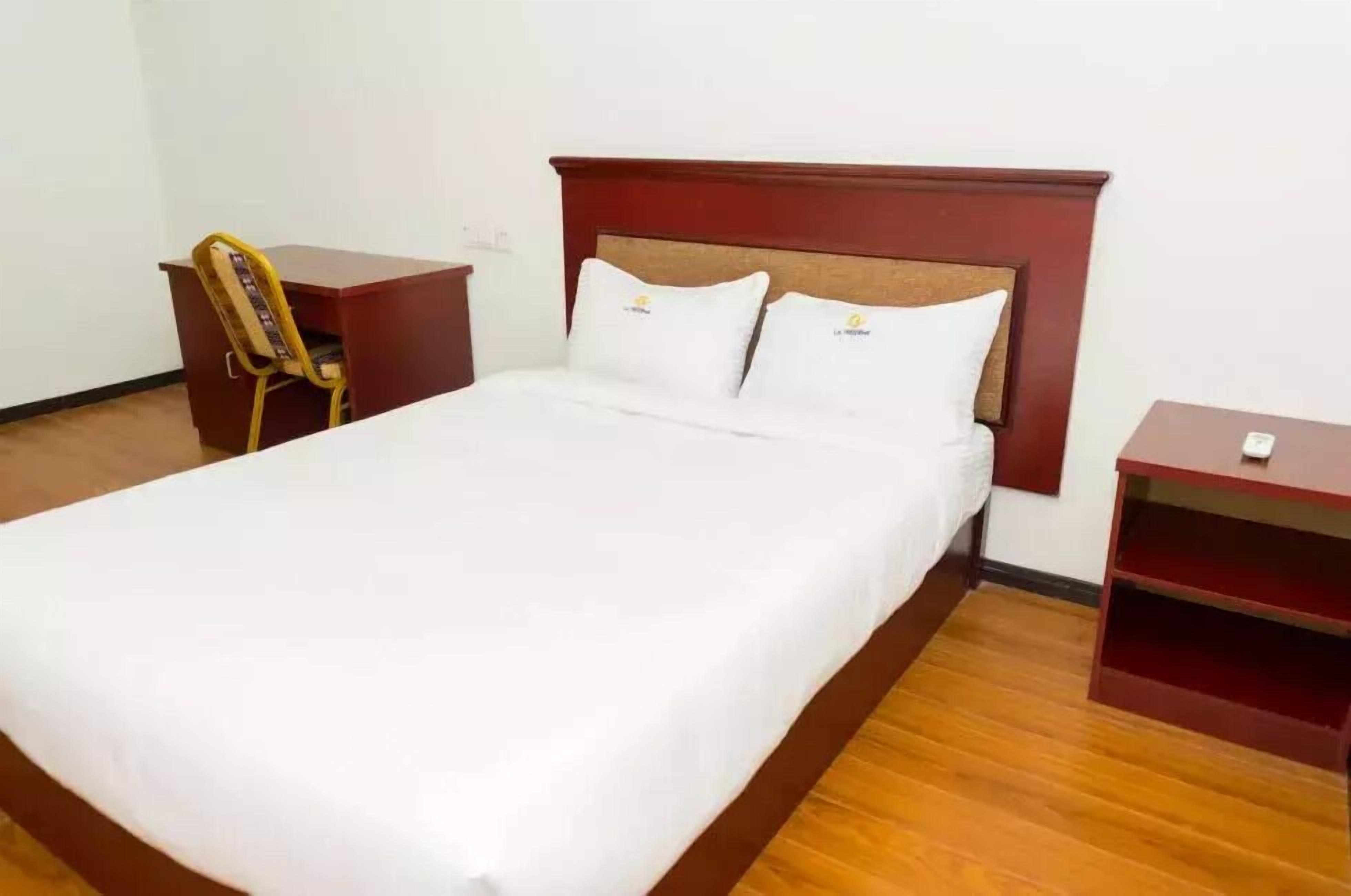 A neatly made bed with white linens and two pillows, a wooden nightstand with a lamp, and a small wooden shelf unit.