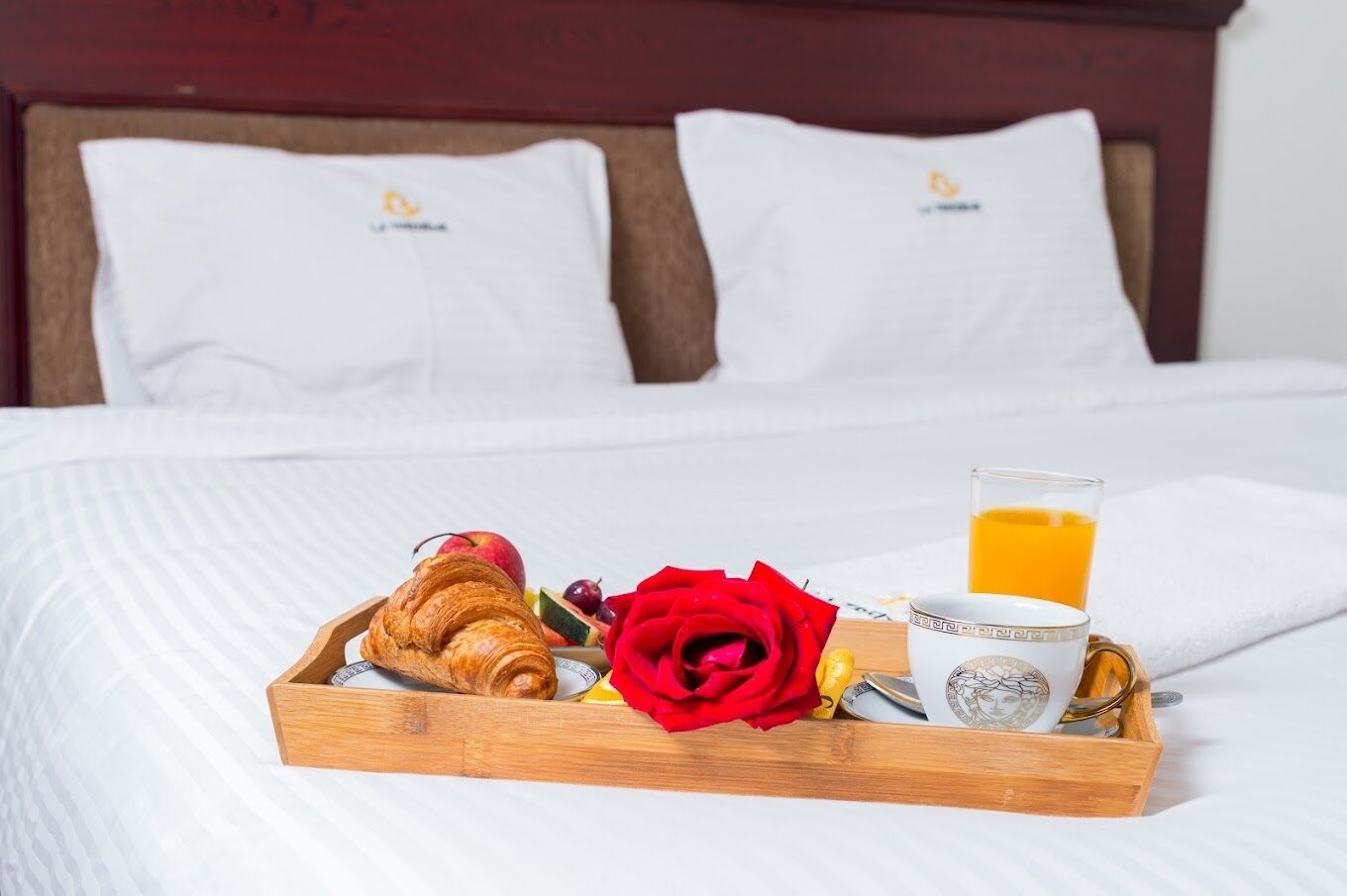Un lit bien fait, avec du linge de lit blanc et des oreillers, un plateau en bois sur lequel on trouve des croissants, des fruits, une rose rouge, un verre de jus d’orange et une tasse au décor raffiné.