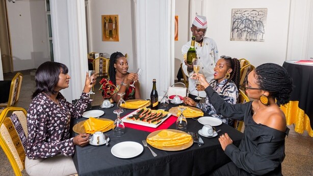 Un groupe de personnes est assis autour d’une table, où se trouvent de la nourriture et des boissons, dans une pièce aux murs blancs et ornée d’un lustre.