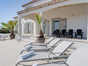 Een zonnig terras met witte lounge stoelen, een tafel met stoelen en potplanten.