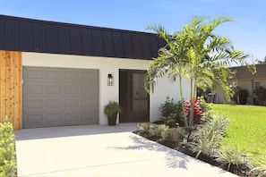 Une maison moderne avec un garage, une porte d’entrée, un petit jardin avec des plantes et des fleurs, et un ciel dégagé.