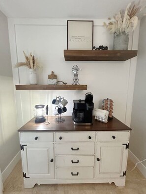 Un plan de travail de cuisine avec une machine à café, une bouilloire et un porte-tasses. Au-dessus du plan de travail, on trouve deux étagères en bois ornées d’objets décoratifs.