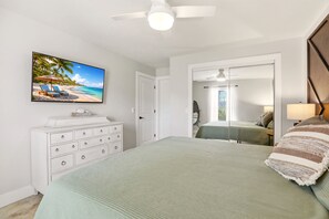 Un dormitorio con cama, cómoda, ventilador de techo, televisor en la pared y puerta que da a otra habitación.