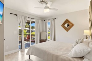 Een slaapkamer met een bed, een plafondventilator, uitzicht door de glazen deur en een tafeltje met een lamp.