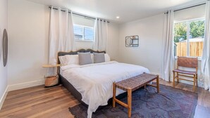 Une chambre à coucher avec un grand lit, un banc en bois, une chaise, une table de chevet et une fenêtre avec des rideaux.