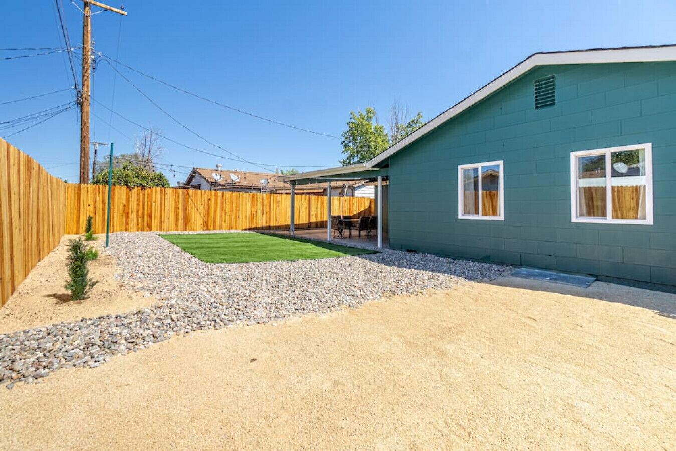 Un jardin arrière d’une maison, avec une pelouse verdoyante, un chemin de gravier, une clôture en bois et une maison dont l’extérieur est vert et les bordures blanches.