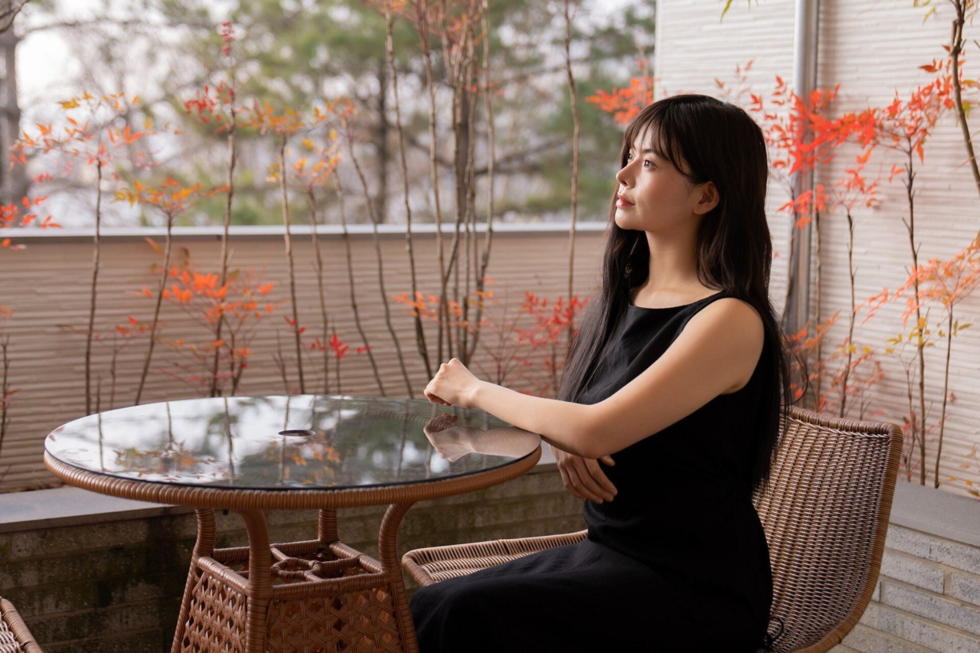 A person seated at a round glass table with a wicker chair, outdoors with a view of trees and a building in the background.
