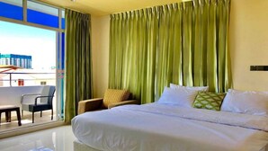 A hotel room with a large bed, a chair, a small table, and a view of buildings through a window. - Contento Retreat (Hulhumalé)
