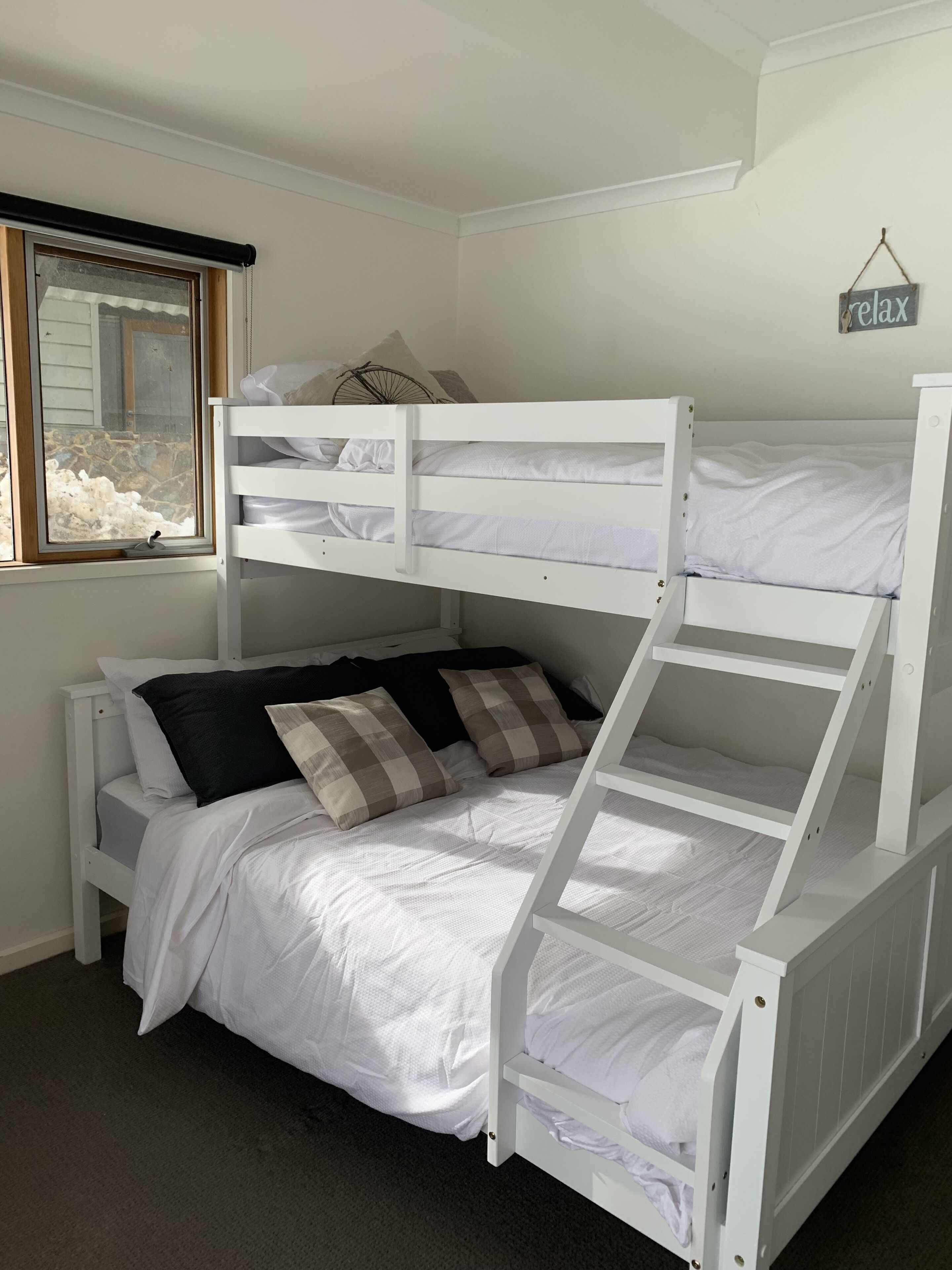 Una cama litera con ropa de cama blanca y almohadas a cuadros, una ventana con vistas a un muro de piedra y un cartel que dice 'relax'.