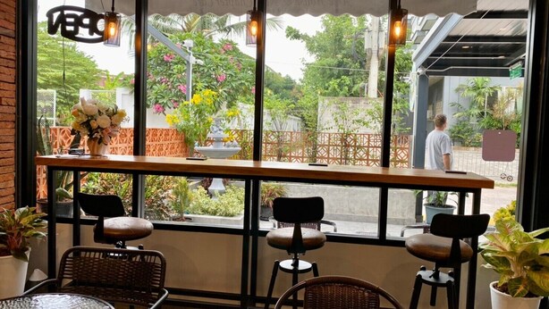 An outdoor seating area with wicker chairs and tables, a wooden bar, and a view of a garden with plants and flowers. - Aree House Poshtel (Pattaya)