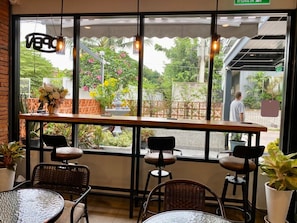 An outdoor seating area with wicker chairs and tables, a wooden bar, and a view of a garden with plants and flowers. - Aree House Poshtel (Pattaya)