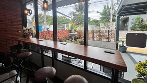 A bar area with a long wooden counter, multiple chairs, and a view of a garden through large windows. - Aree House Poshtel (Pattaya)