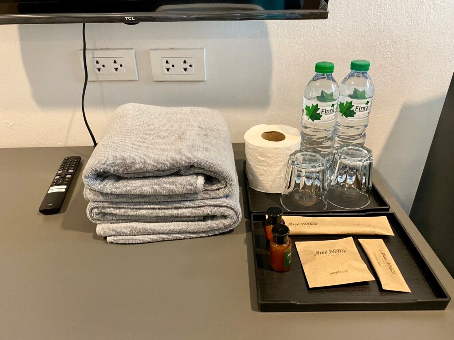 Une chambre d’hôtel avec une serviette soigneusement pliée, une télécommande, un rouleau de papier toilette, un plateau avec une bouteille de shampoing, une bouteille de gel douche, un verre d’eau et un verre de jus de fruits.