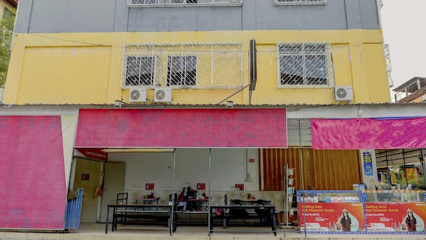 Un edificio de dos pisos con fachada amarilla y gris, un toldo rojo sobre la planta baja, y una tienda con toldo rosa a la derecha.