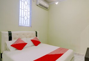 A bed with a white headboard and two pillows, one red and one white, in a room with a window and an air conditioning unit. - Hotel O Backpaker Seraya (Batam)