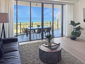 Uma sala de estar moderna com uma grande porta de vidro deslizante que dá para uma varanda com vista para o oceano e um prédio distante.