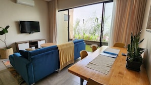 A modern living room with a blue sofa, a wooden dining table, and a TV mounted on the wall. - Instahomes by Tru - Garden Suites (Blantyre)